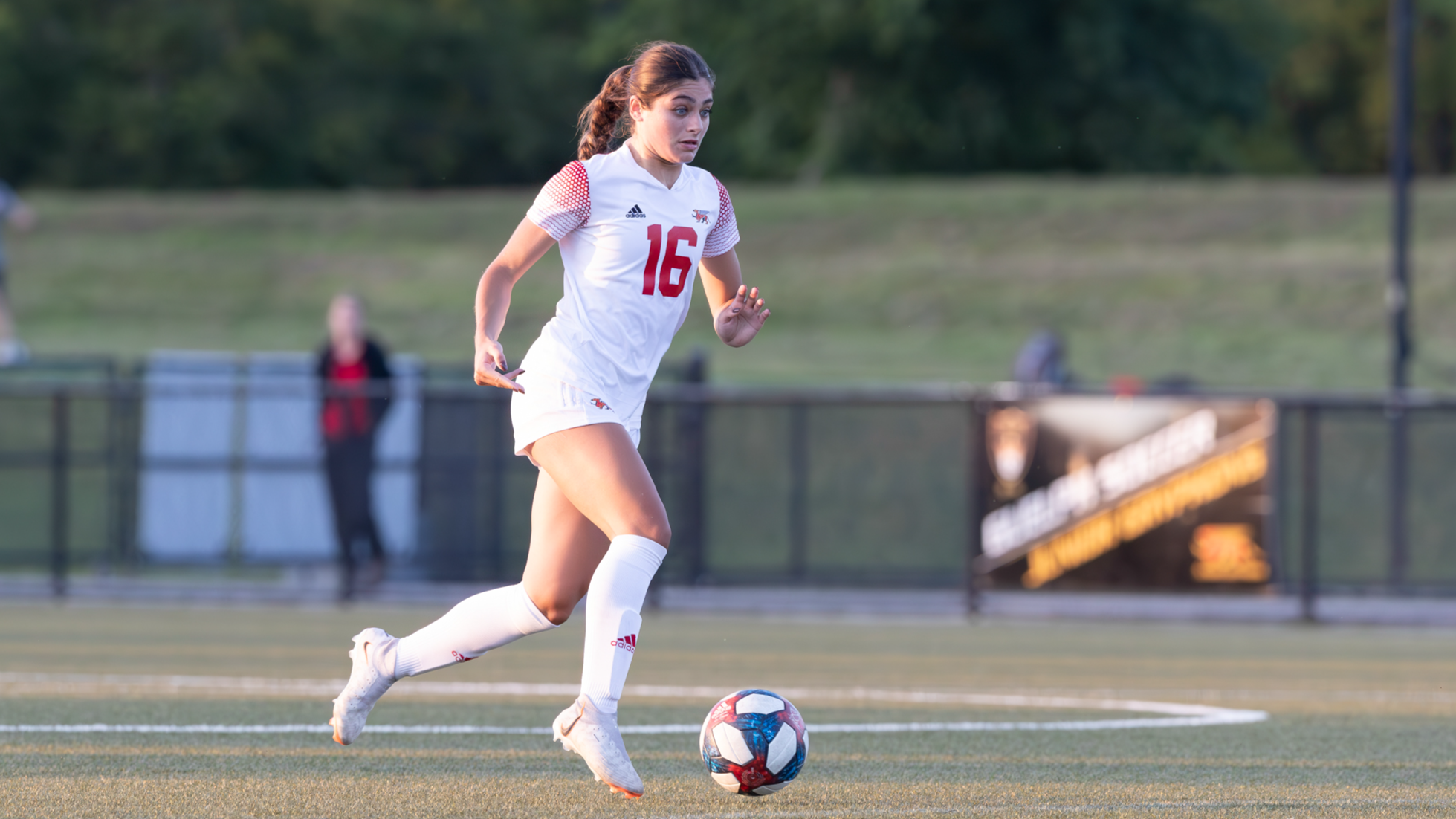 Image of woman in white soccer jersey and shorts with red numbers and details running on a soccer pitch kicking a soccer ball
